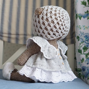 May include: A crocheted teddy bear wearing a white dress with a lace trim and a crocheted hat. The bear is sitting on a blue surface.