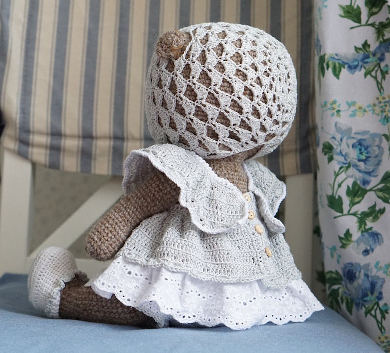 May include: A crocheted teddy bear wearing a white dress with a lace trim and a crocheted hat. The bear is sitting on a blue surface.