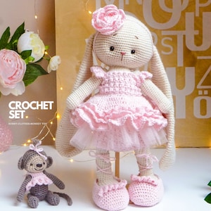 Może przedstawiać: Zestaw do szydełkowania z dużą, kremową lalką królika w różowej sukience i mniejszą szarą lalką małpy. Królik ma długie uszy i kwiat na głowie. Widoczny jest tekst "CROCHET SET".