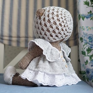 May include: A crocheted teddy bear wearing a white crocheted dress with a lace trim and a matching crocheted hat.