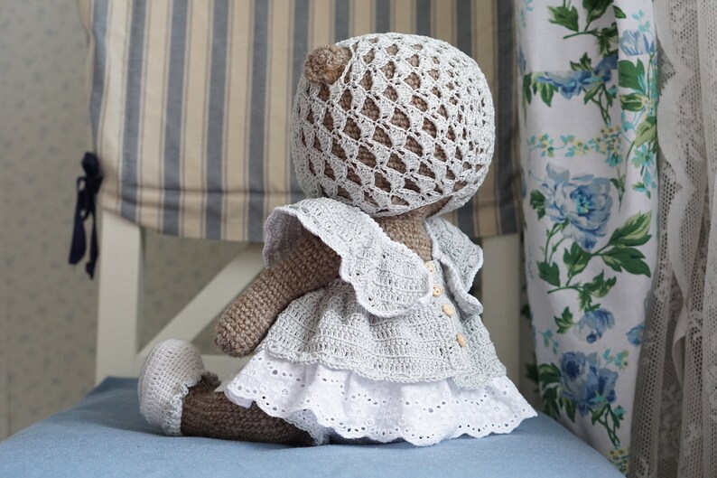 May include: A crocheted teddy bear wearing a white crocheted dress with a lace trim and a matching crocheted hat.