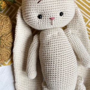 May include: A crocheted cream-colored bunny with long floppy ears. The bunny has black eyes and a pink nose.