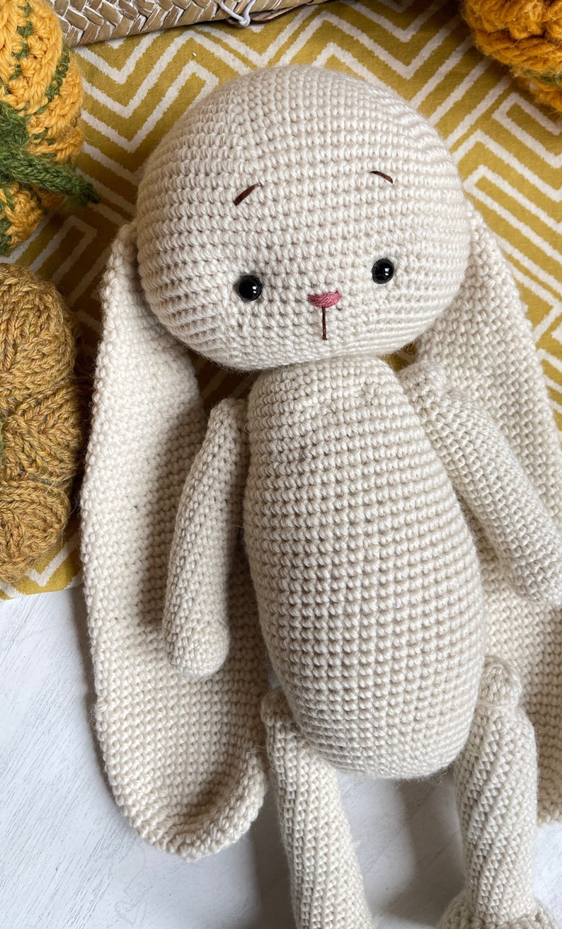 May include: A crocheted cream-colored bunny with long floppy ears. The bunny has black eyes and a pink nose.