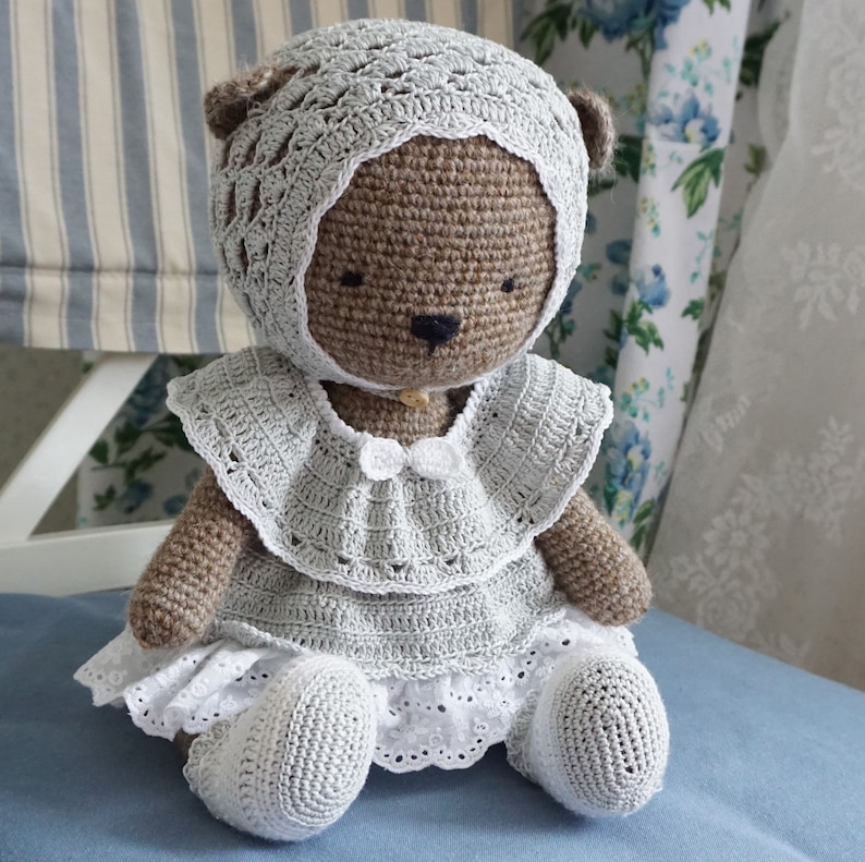 May include: A crocheted teddy bear wearing a white bonnet and a light blue dress with white lace trim. The bear is sitting on a blue surface.