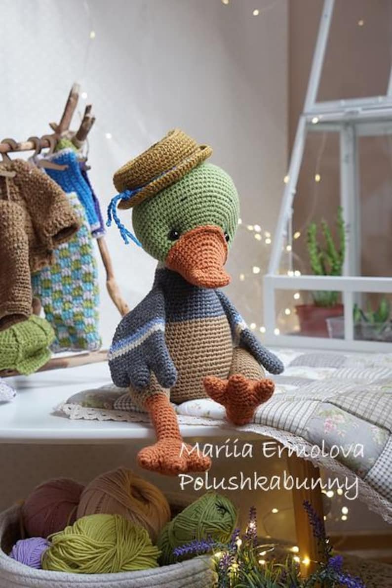 Op de afbeelding: Een handgemaakte gehaakte eendenpop met een groene kop, oranje snavel en voeten en een bruin en blauw lichaam. De eend draagt een kleine bruine hoed en zit op een wit oppervlak. De afbeelding bevat garen en kleine gehaakte kleding.