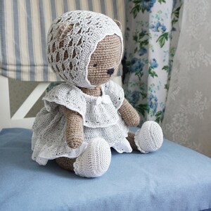 May include: A crocheted teddy bear wearing a white dress and bonnet. The bear is sitting on a blue chair.