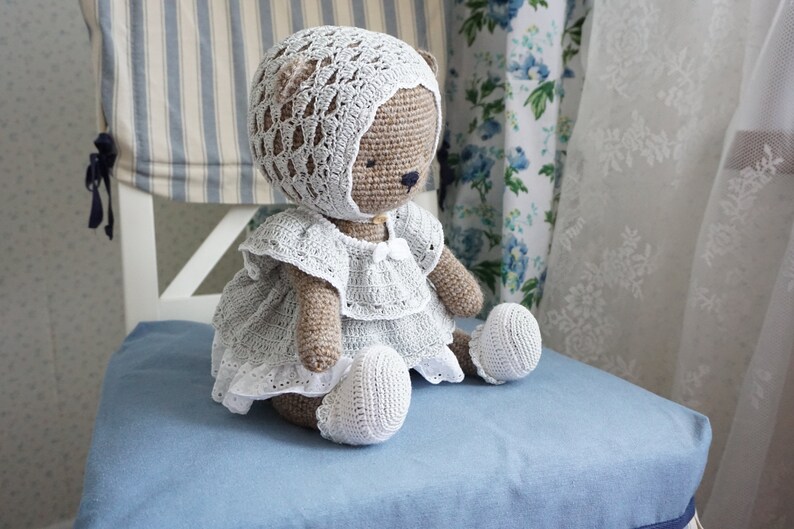 May include: A crocheted teddy bear wearing a white dress and bonnet. The bear is sitting on a blue chair.