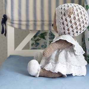 May include: A crocheted teddy bear wearing a white dress and bonnet. The bear is sitting on a blue chair with a striped fabric backing.