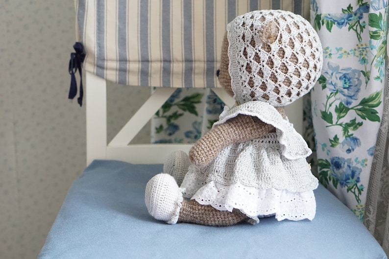 May include: A crocheted teddy bear wearing a white dress and bonnet. The bear is sitting on a blue chair with a striped fabric backing.