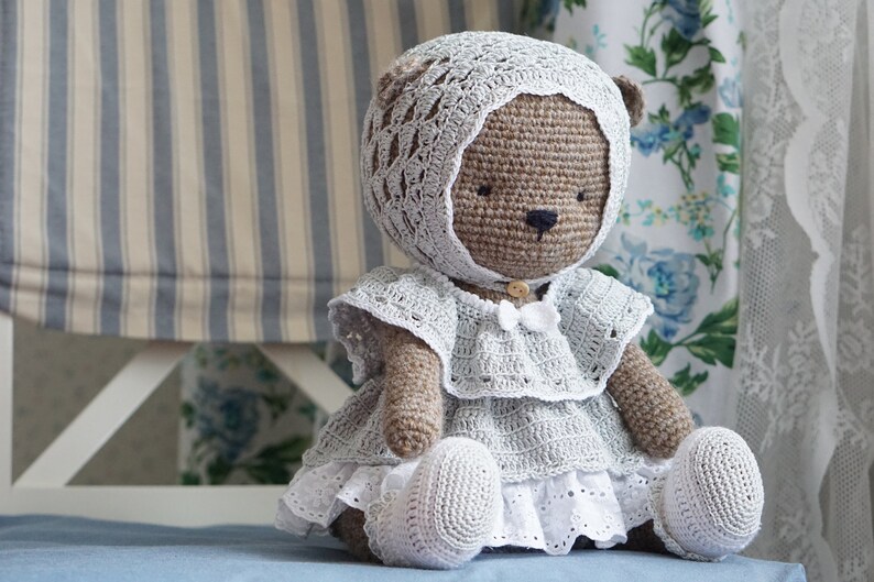 May include: A crocheted teddy bear wearing a white lace dress and a matching bonnet. The bear is sitting on a blue surface.