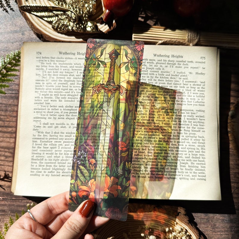 May include: A stained glass bookmark featuring a sword with a golden hilt and a red, green, and yellow floral design. The bookmark is translucent and shows the text "Wuthering Heights" from a book.