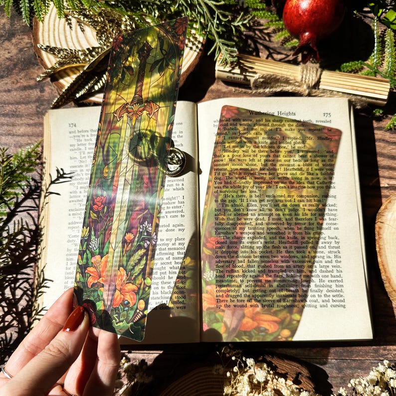 May include: A colourful bookmark featuring a stained glass style illustration of a sword surrounded by flowers. The bookmark is placed on an open book with the text "Wuthering Heights" visible.