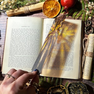 May include: A close-up of a bookmark featuring a stained glass design of a dragonfly with yellow, orange, and brown colours. The bookmark is placed on a book with the text "CHAPTER XXI" visible.