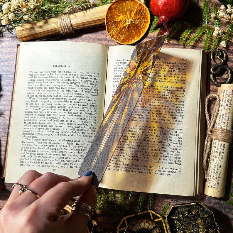 May include: A close-up of a bookmark featuring a stained glass design of a dragonfly with yellow, orange, and brown colours. The bookmark is placed on a book with the text "CHAPTER XXI" visible.