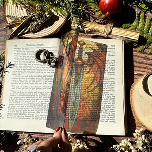 May include: A colourful bookmark featuring a detailed illustration of a dragonfly with a sword in its mouth. The bookmark is made of a translucent material and is placed on a book with the text "Wathering Heights" visible.