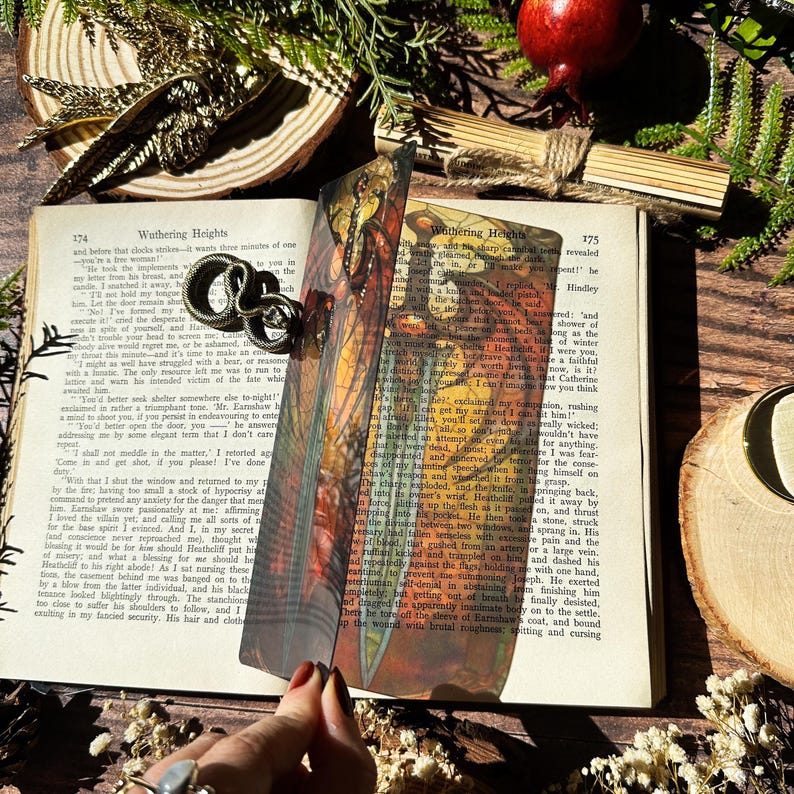 May include: A colourful bookmark featuring a detailed illustration of a dragonfly with a sword in its mouth. The bookmark is made of a translucent material and is placed on a book with the text "Wathering Heights" visible.