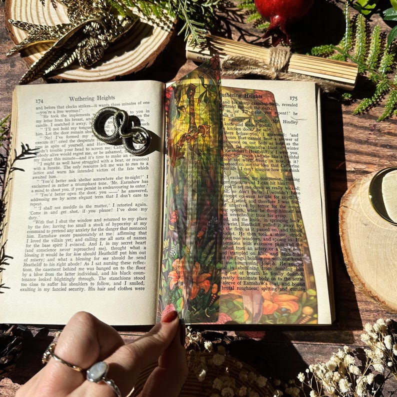 May include: A close-up of a book with a bookmark featuring a colourful floral design. The book is open to a page with the title "Wuthering Heights" and text from the novel. The bookmark is made of glass and has a metallic finish.