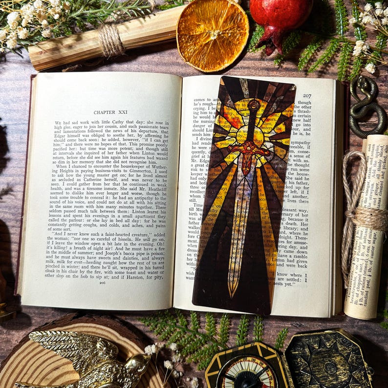 May include: A stained glass bookmark depicting a golden sword with a cross hilt, surrounded by a bright yellow sunburst. The sword is set against a dark brown background.