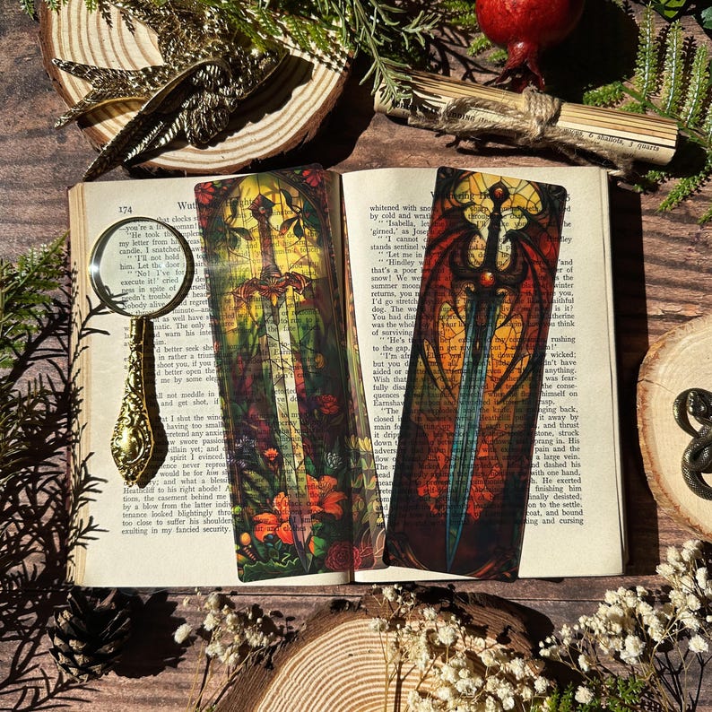May include: Two colourful bookmarks featuring stained glass style illustrations of swords. The bookmarks are set against a background of a book with text.