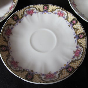 May include: A white porcelain saucer with a scalloped edge and a floral design in shades of pink, purple, and gold. The design features a chequered pattern, floral motifs, and ribbon accents.