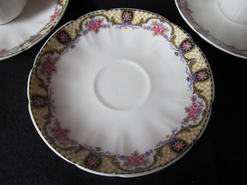 May include: A white porcelain saucer with a scalloped edge and a floral design in shades of pink, purple, and gold. The design features a chequered pattern, floral motifs, and ribbon accents.