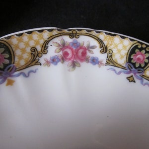 May include: A white ceramic saucer with a gold and black floral design. The design features a repeating pattern of small flowers and leaves, with a larger floral motif in the centre. The saucer has a scalloped edge.