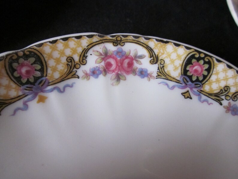 May include: A white ceramic saucer with a gold and black floral design. The design features a repeating pattern of small flowers and leaves, with a larger floral motif in the centre. The saucer has a scalloped edge.