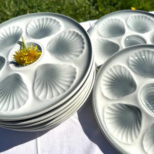 Puede incluir: Una pila de platos de cerámica azul claro para ostras con hendiduras en forma de concha. Un diente de león amarillo descansa en el centro de un plato. Los platos están sobre una superficie blanca, con un fondo verde borroso.