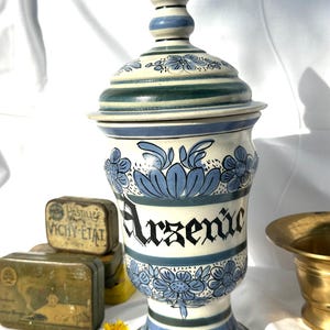Peut inclure: Un pot d'apothicaire en céramique avec couvercle, présentant une base crème avec des motifs floraux bleus et des rayures horizontales. Le mot "Arsenic" est inscrit en lettres gothiques noires sur le devant. Le pot a une base évasée.