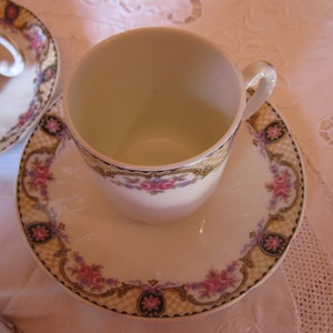 May include: A white porcelain teacup and saucer with a floral pattern in shades of pink, purple, and yellow. The saucer has a gold trim and a chequered pattern.