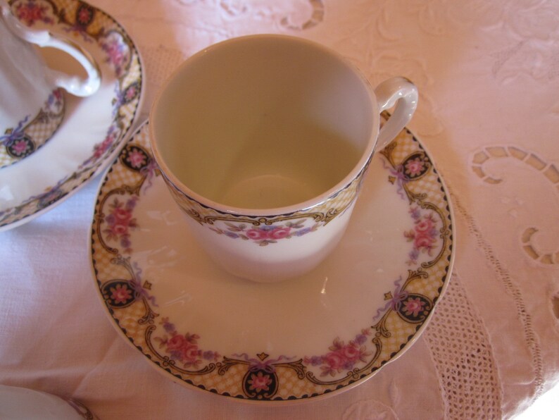 May include: A white porcelain teacup and saucer with a floral pattern in shades of pink, purple, and yellow. The saucer has a gold trim and a chequered pattern.