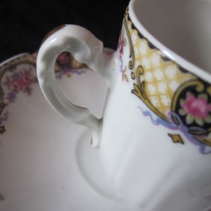 May include: A white porcelain teacup and saucer with a gold and black floral design. The cup has a delicate handle and the saucer has a scalloped edge.