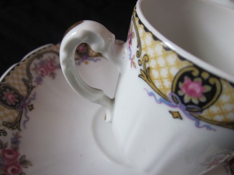 May include: A white porcelain teacup and saucer with a gold and black floral design. The cup has a delicate handle and the saucer has a scalloped edge.