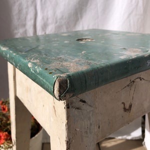 Könnte beinhalten: Ein vintage Holzhocker mit einer grün lackierten Oberseite und weiß lackierten Beinen. Die Farbe ist abgeblättert und abgenutzt, was dem Hocker ein rustikales Aussehen verleiht.