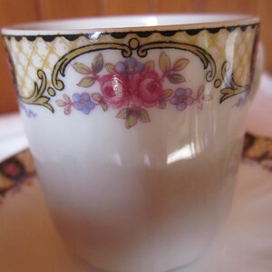 May include: A white porcelain teacup with a gold and black floral design. The cup features a delicate floral pattern with pink, blue, and yellow flowers.