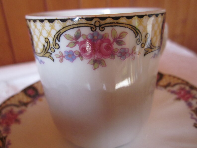 May include: A white porcelain teacup with a gold and black floral design. The cup features a delicate floral pattern with pink, blue, and yellow flowers.