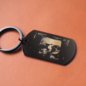 May include: Black metal keyring with a dog tag-shaped pendant. The pendant features a sonogram image in a lighter tone against a black backdrop. The keyring is a dark grey colour. The background is a solid, reddish-brown colour.