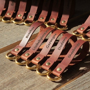 May include: Several brown leather keyrings with gold-coloured rings and rivets, each engraved with a name. The keyrings are displayed on a wooden surface, close-up shot.