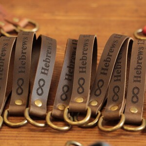 May include: Brown leather keychains with the text "Hebrews 13" and an infinity symbol. The keychains have gold-coloured metal rings and studs. The background is a wooden surface.
