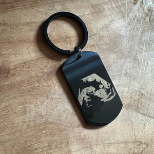May include: A black metal dog tag keyring featuring a laser-engraved ultrasound image on the front surface. The rectangular tag has rounded corners and is attached to a circular black metal split ring, creating a sleek and functional accessory.