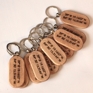 May include: A collection of oval-shaped wooden keychains, each featuring a silver metal split ring. The light-toned wood surfaces are engraved with the black text 39° 8' 19.1652" N 84° 30' 15.3144" W, displaying precise geographic coordinates.
