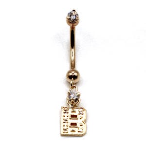 May include: Gold-colored belly button ring with a curved barbell design. The ring features a small, clear gemstone at the top and a dangling charm shaped like the letter "B" with a small gemstone. The jewelry is set against a white background.
