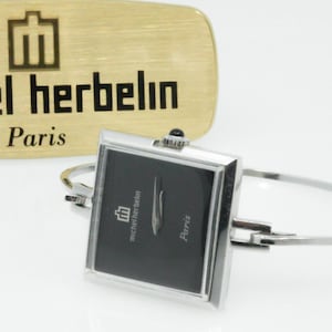 May include: A silver-tone bangle watch with a square black face featuring the text michel herbelin Paris. Behind the watch is a gold-tone rectangular plaque with the same brand logo and text engraved in black.