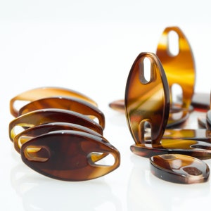 Könnte beinhalten: Eine Gruppe von braunen Schildpatt-Haarklammern mit einer einzigartigen ovalen Form und zwei Löchern in der Mitte. Die Klammern sind in einem zufälligen Muster auf einer weißen Oberfläche angeordnet.