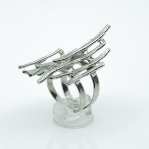 Könnte beinhalten: Ein silberfarbener Ring mit abstraktem Design. Der Ring hat mehrere Bänder und einen oberen Bereich mit einer Reihe paralleler, strukturierter Stäbe. Der Ring wird auf einem transparenten Ständer vor weißem Hintergrund präsentiert.