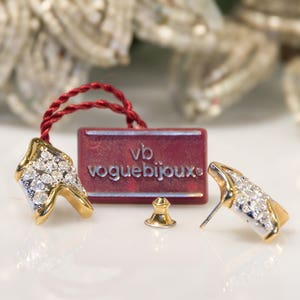 Könnte beinhalten: Ein Paar goldene Ohrringe mit klaren Kristallsteinen, eine rote Schachtel mit dem Text "vb voguebijoux" und ein goldener Ohrringverschluss.