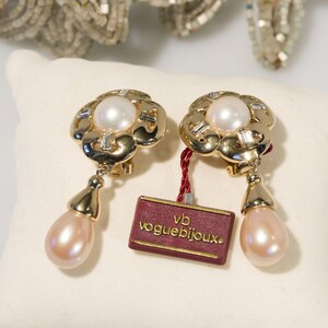 May include: Gold-toned clip-on earrings with a floral design, featuring a central pearl and baguette-cut accents. Each earring has a teardrop-shaped, pink pearl drop. A tag reads "vogue bijoux."