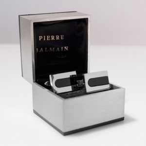 Può includere: Una scatola per gemelli d'argento con interno nero. La scatola è aperta e mostra due gemelli d'argento con inserti neri. La scatola ha la scritta "PIERRE BALMAIN PARIS" sul coperchio.