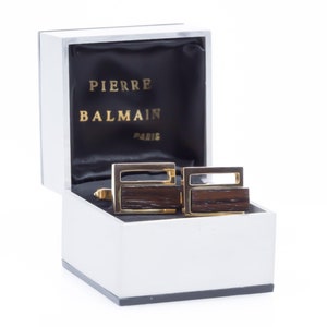 May include: A pair of gold cufflinks with wood inlays in a silver box. The box has the text "PIERRE BALMAIN PARIS" on the inside.