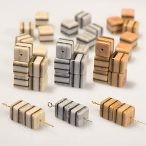 May include: A collection of striped, cube-shaped beads in shades of brown, beige, and grey. The beads are arranged in stacks of three, with some beads strung on thin wires.